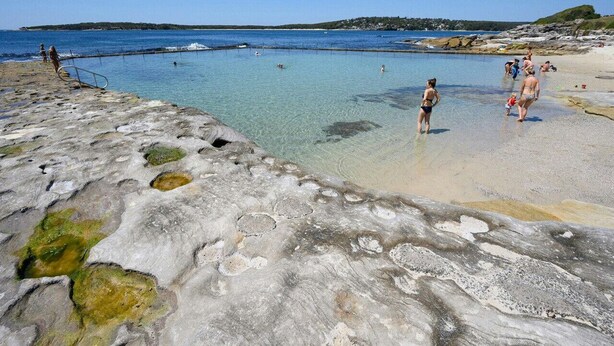Una spiaggia rocciosa con acque cristalline, persone che nuotano e una costa lontana.