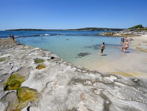 A rocky beach with clear water, people swimming, and a distant shoreline. - Airy Cronulla beach escape 2 bed (Cronulla)