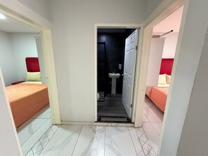 Un pasillo estrecho que conduce a un baño con lavamanos e inodoro. A ambos lados del pasillo hay dos camas, una con cabecera roja y la otra con cabecera blanca.