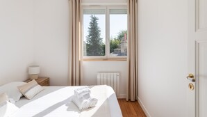 Un dormitorio ordenado con una cama grande, una mesita de noche con lámpara, una ventana con cortinas y vistas a árboles y edificios.