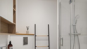 Un baño moderno con lavamanos, inodoro y ducha blancos. El suelo presenta un diseño y hay un estante de madera sobre el lavamanos.