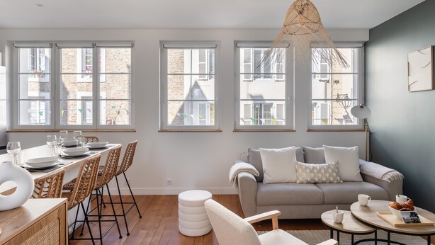 Una sala de estar moderna con sofá, mesa de comedor y sillas. Amplias ventanas con contraventanas, una lámpara colgante y vistas a edificios.