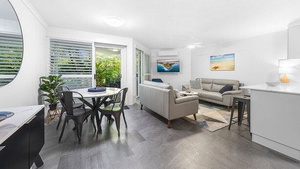 En moderne stue med sofa, spisebord med stole, et køkkenområde med vask og et stort vindue med persienner.
