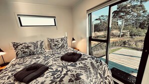 Une chambre avec un grand lit, une literie à motifs floraux, une fenêtre donnant sur une terrasse et des arbres, et une prise électrique sur le mur.