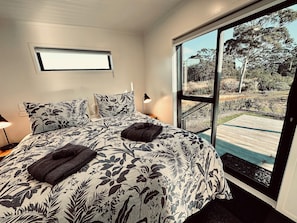 Quarto com cama grande, roupa de cama floral, janela com vista para um deck e árvores, e uma tomada na parede.