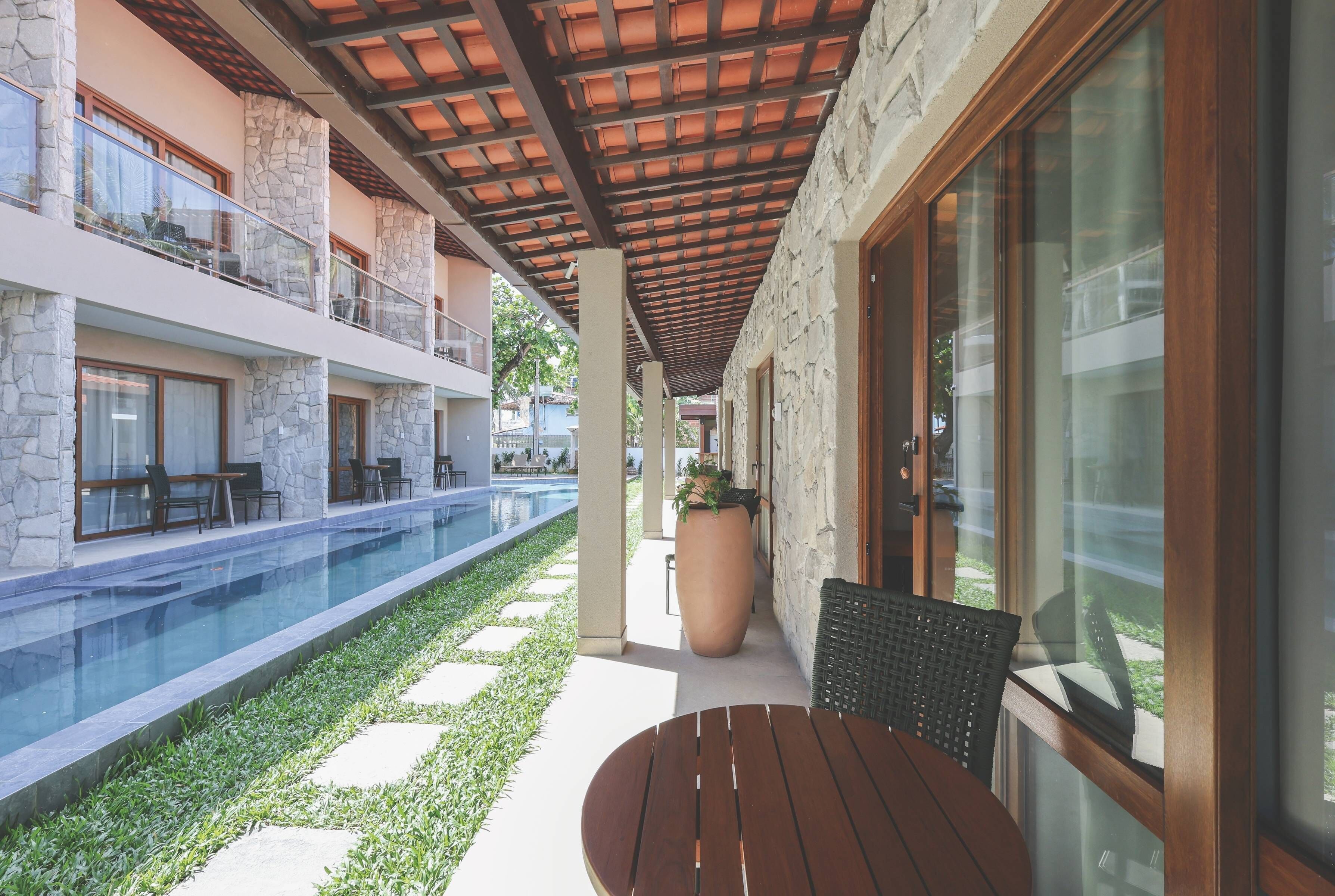 A poolside area with a wooden table and chairs, stone pillars, and a glass-enclosed balcony.