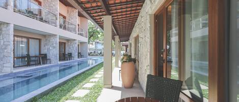 A poolside area with a wooden table and chairs, stone pillars, and a glass-enclosed balcony.