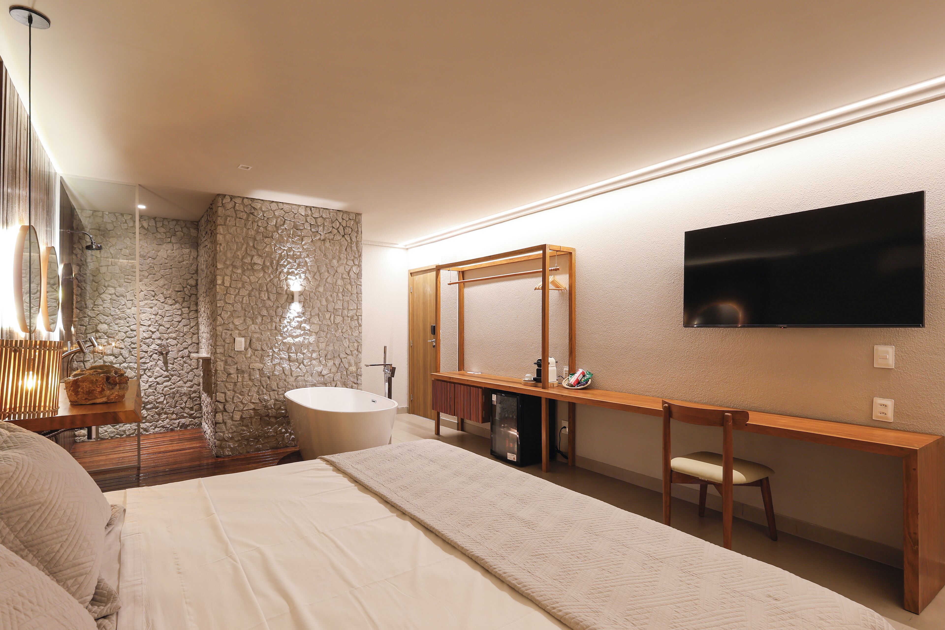 A modern bedroom with a large bed, a wooden desk, a television, a bathroom with a stone wall, and a shower.
