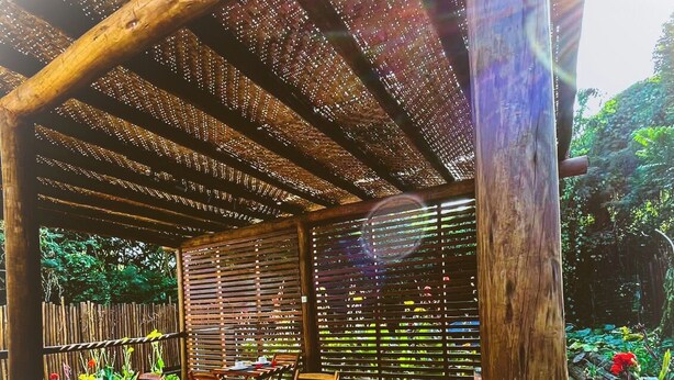 Une terrasse extérieure en bois, dotée de tables et de chaises, entourée d’une clôture en bambou et d’une végétation luxuriante.