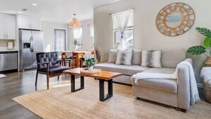 Un salon moderne avec un canapé, un fauteuil et une table basse. On y trouve également un coin repas avec une table et des chaises. Une cuisine équipée d’un réfrigérateur et de placards est visible au fond.