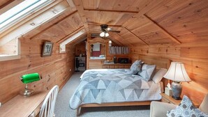 A cozy wooden cabin bedroom with a sloped ceiling, a bed with patterned bedding, a desk with a green lamp, and a view of the kitchen through a skylight.