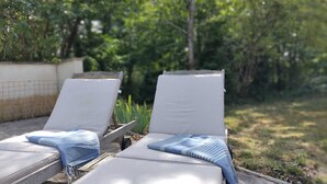 Zwei Liegen mit weißen Kissen und blau gestreiften Handtüchern, draußen aufgestellt, mit Garten und Bäumen im Hintergrund.