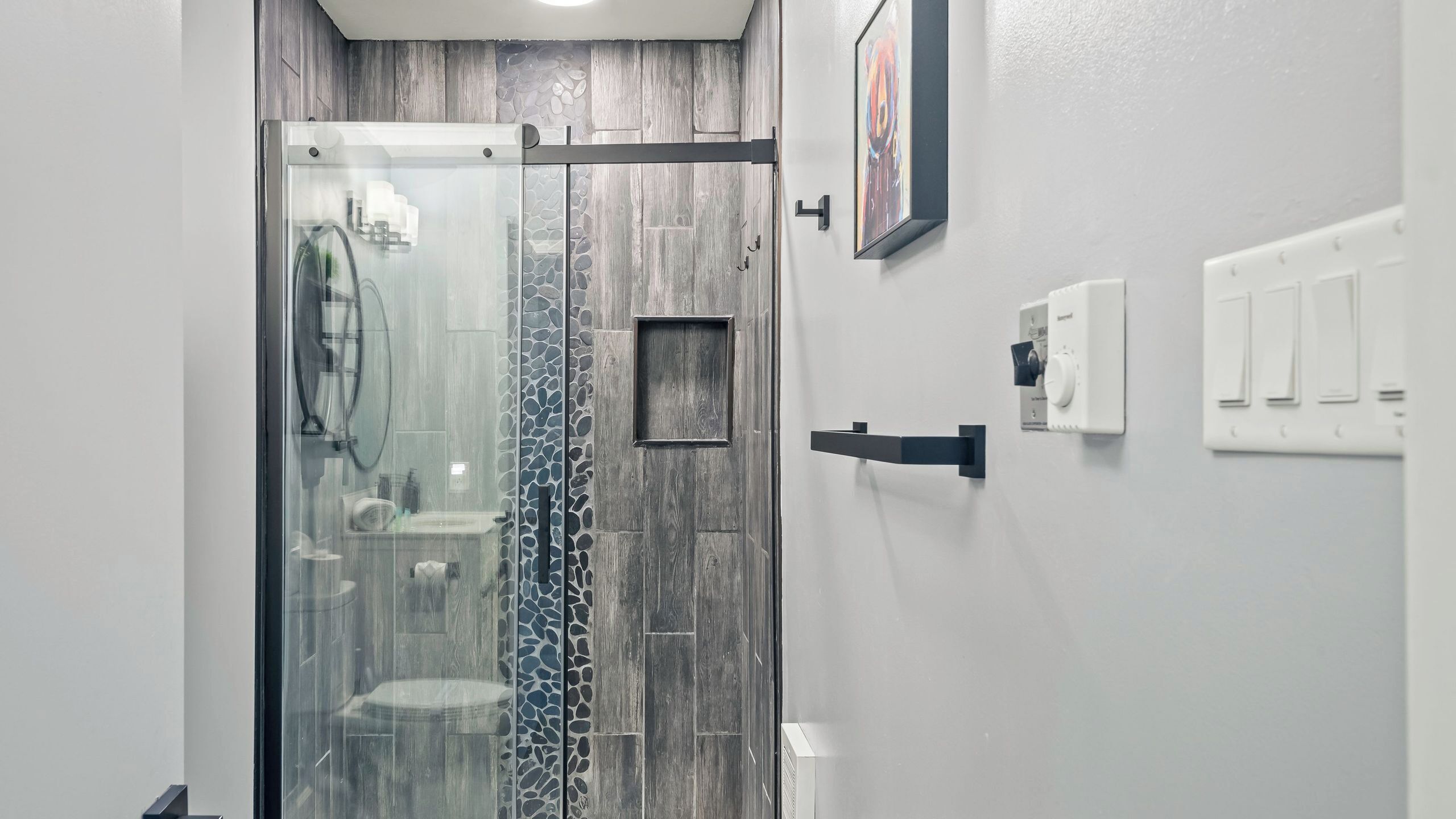 Un baño moderno con cabina de ducha de vidrio, pared con diseño de piedra, un cuadro colgado y un interruptor de luz en la pared.
