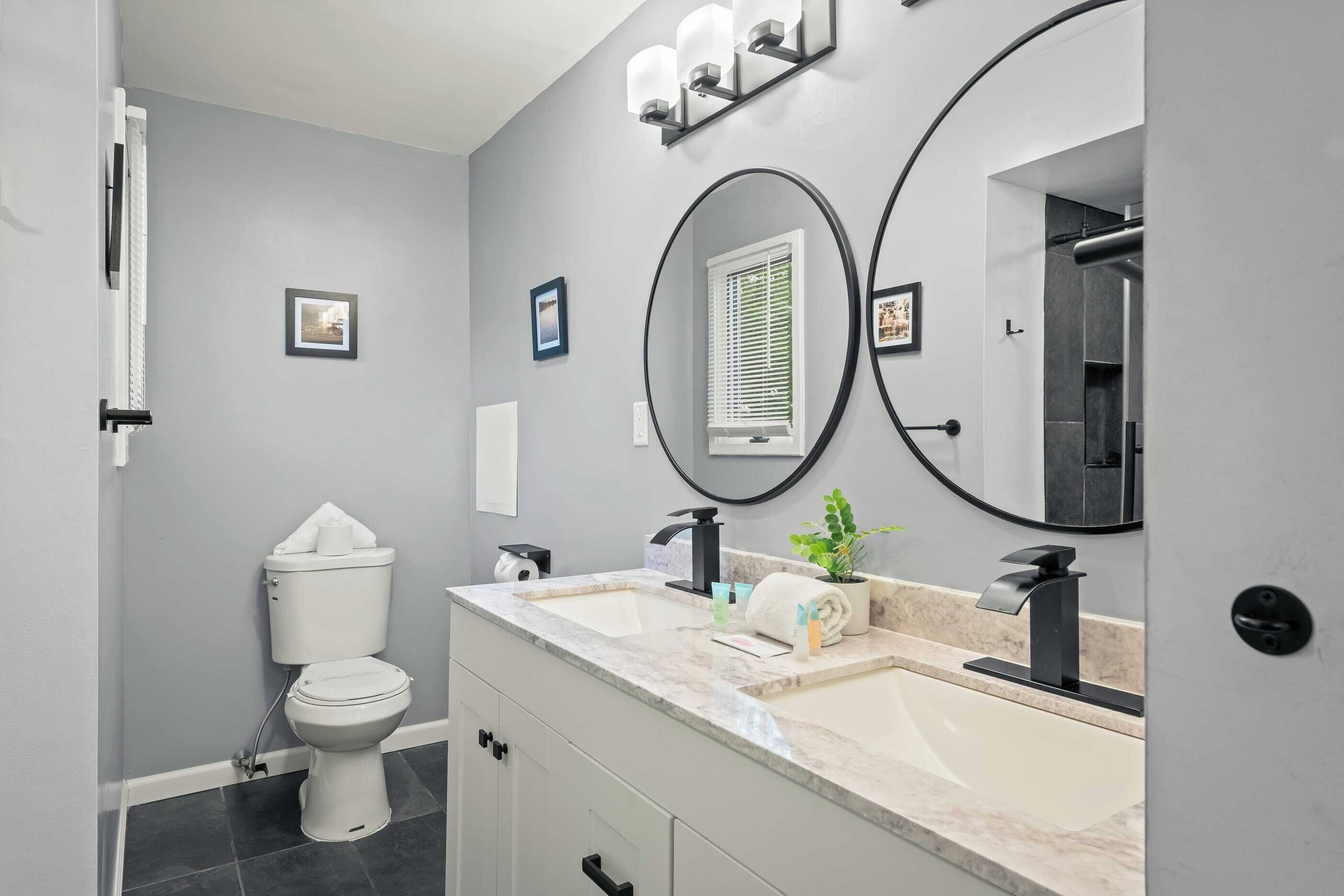 A modern bathroom with a toilet, two sinks, a mirror, and a window with blinds.