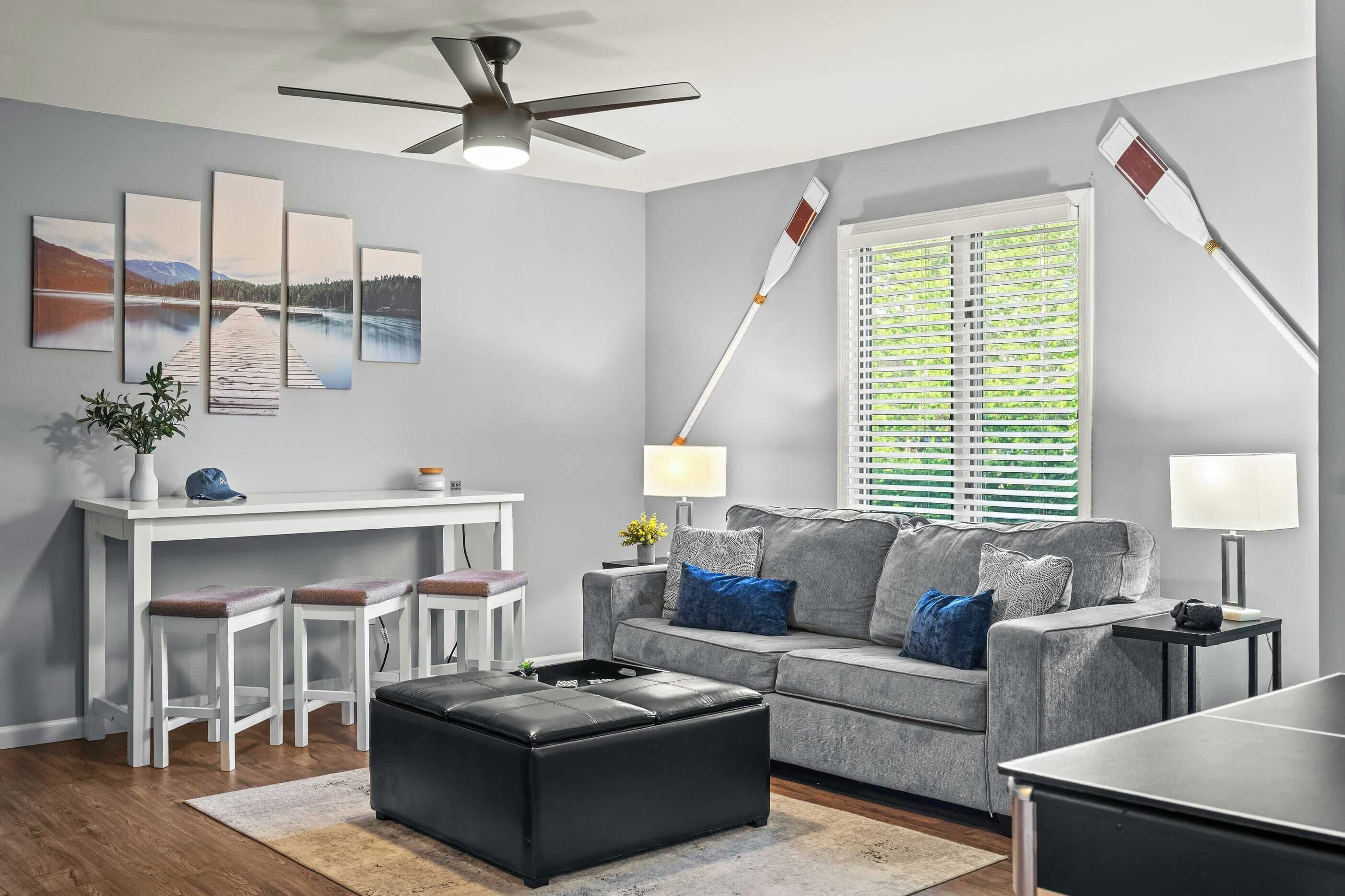 A modern living room with a grey sofa, a black ottoman, a white desk with stools, a ceiling fan, and a large window with blinds.