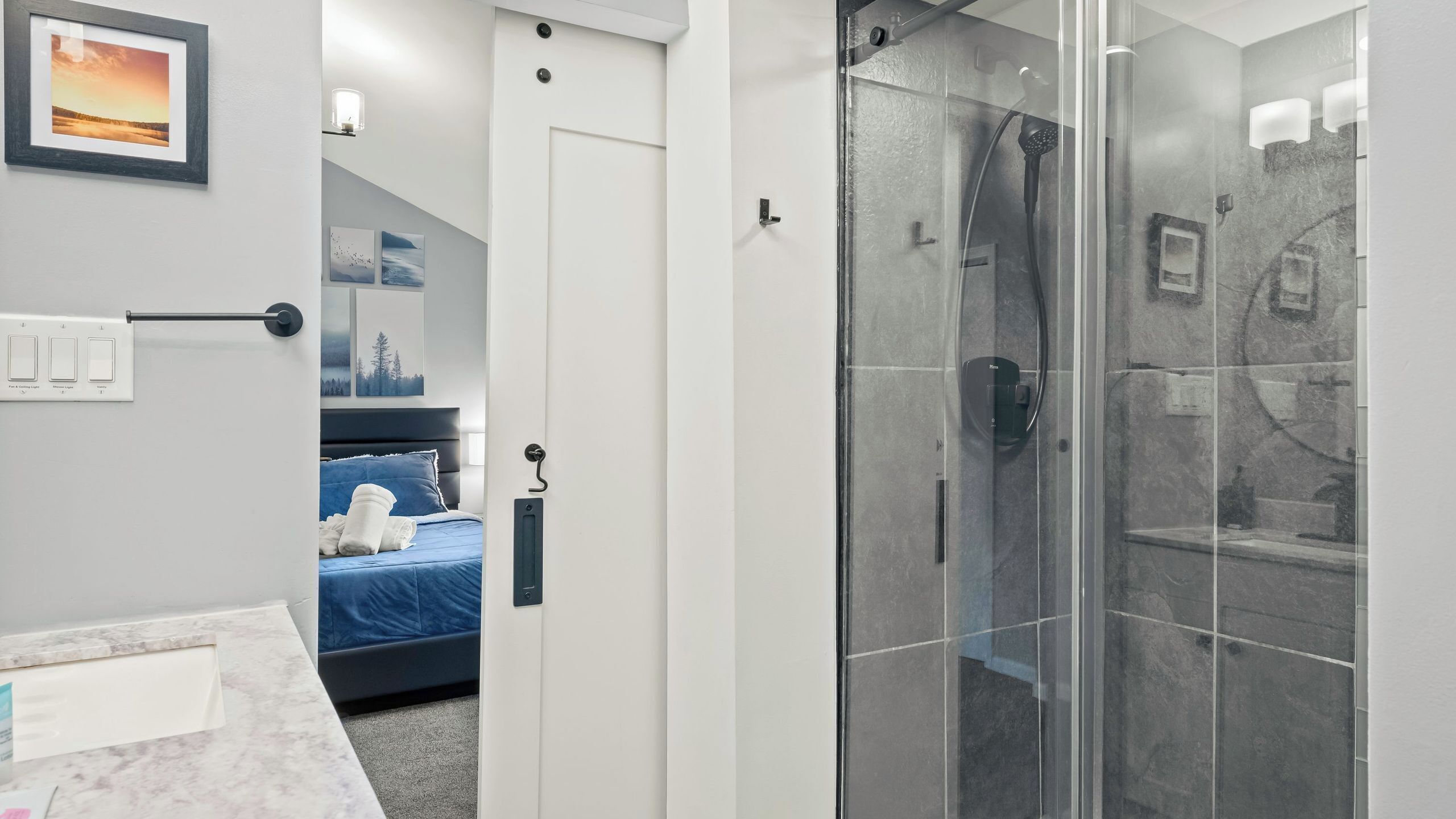 A modern bathroom with a glass shower enclosure, a bed with a blue headboard, and a wall-mounted light fixture.