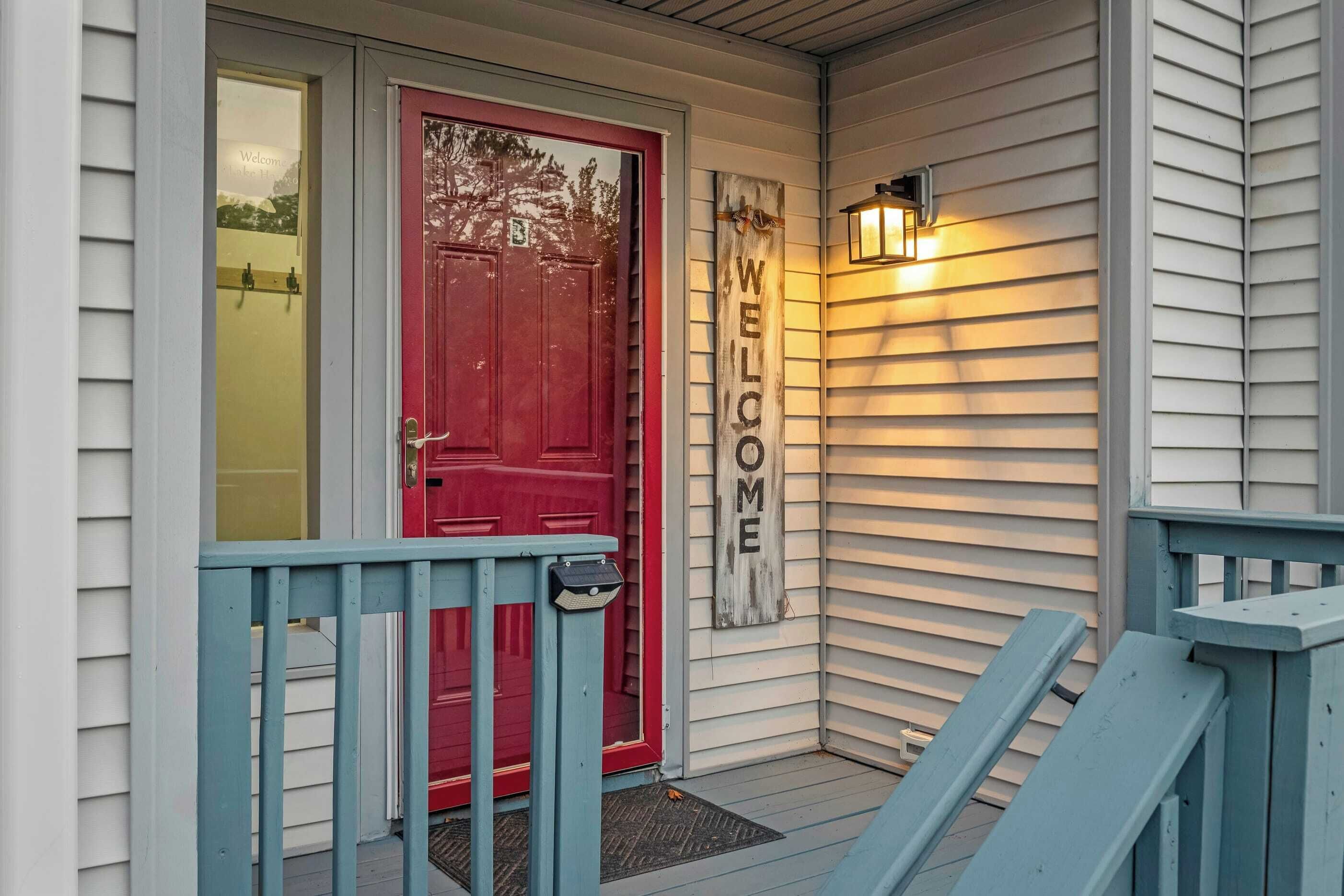 A red door with a 'WELCOME' sign, a mailbox, and a bench on a porch with white siding.