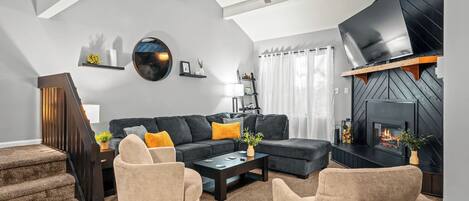 A modern living room with a sectional sofa, two armchairs, a coffee table, a fireplace, a TV, and a staircase.