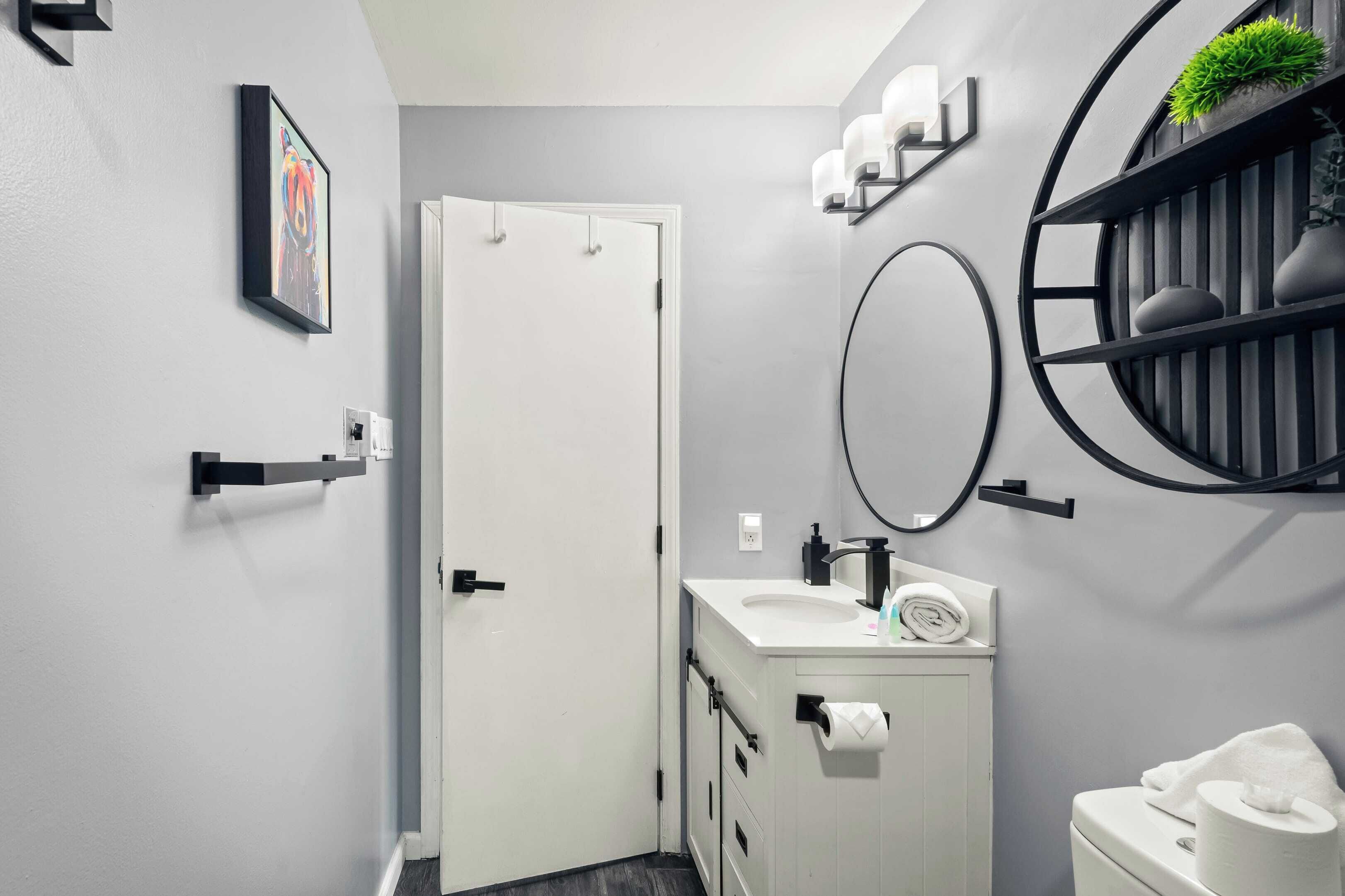 A modern bathroom with a white sink, toilet, and a door. There is a framed picture on the wall and a round mirror with shelves above it.