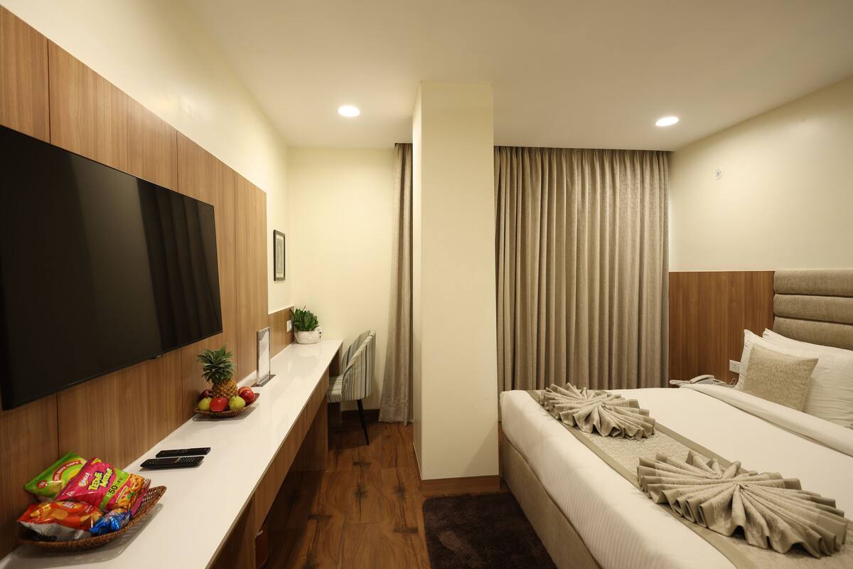 A modern hotel room with a flat-screen TV, a wooden desk with snacks, a bed with decorative pillows, and a large window with curtains.