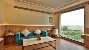 A modern living room with a teal sofa, a coffee table, and a large window offering a view of a green landscape. - Rukman Hotels Jaipur (Jaipur)