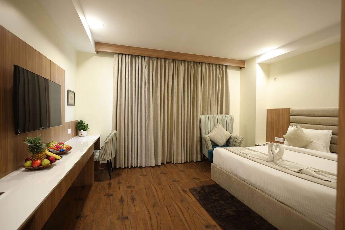 A hotel room with a wooden floor, a bed with a white headboard, a bedside table with a fruit basket, a television mounted on the wall, and a window with curtains.