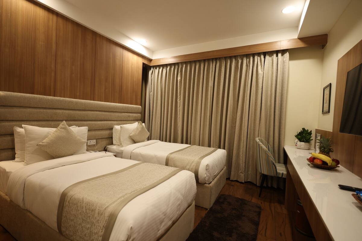 A hotel room with two beds, a desk with a fruit bowl, a chair, and a large window with curtains.