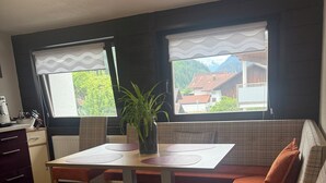 Un coin repas avec une table et des bancs, une plante en pot et une vue sur les maisons à travers les fenêtres.