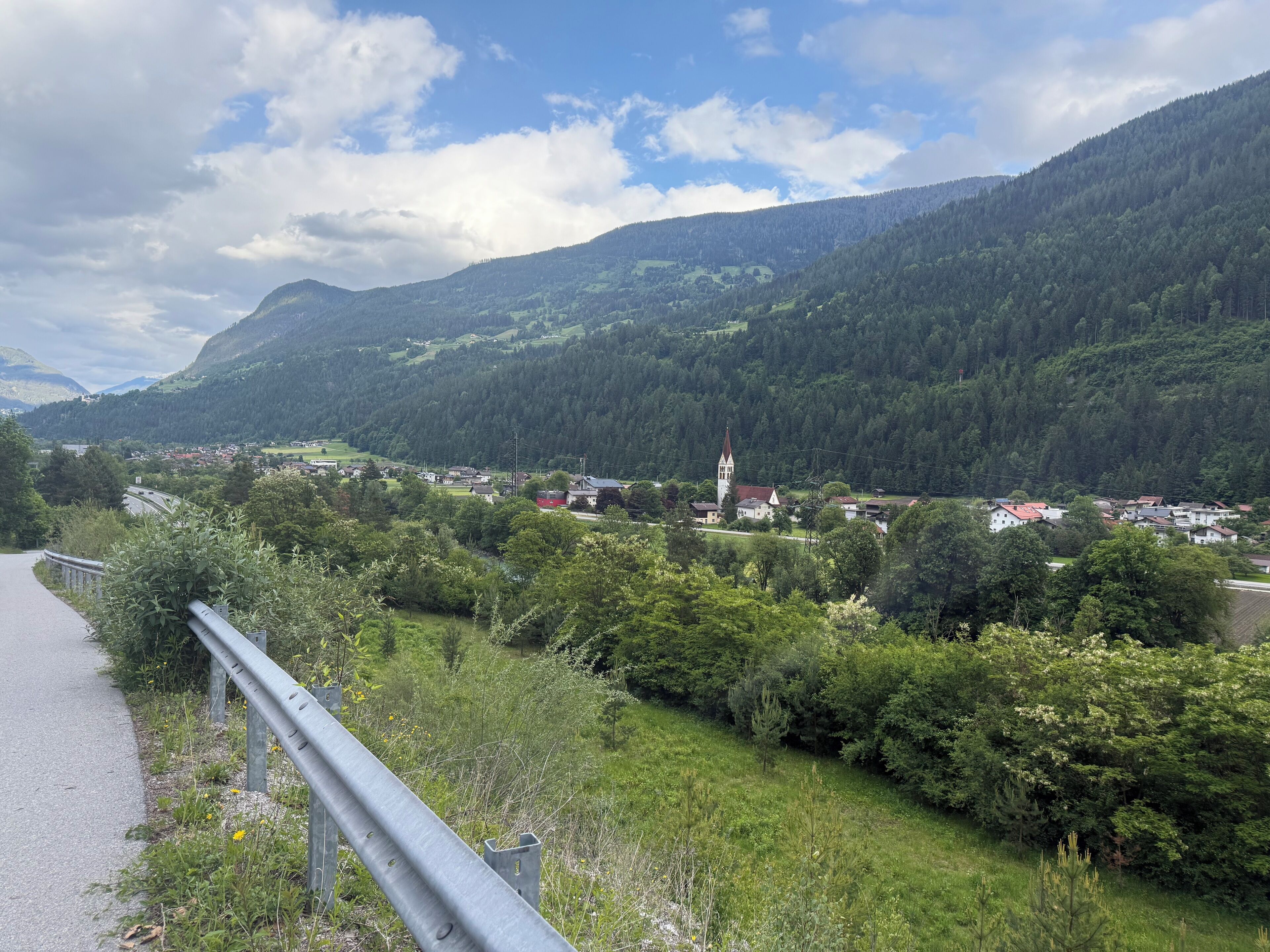 A scenic mountain landscape with a village, a church with a tall steeple, and a road with a guardrail.