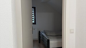 Un couloir étroit mène à une chambre à coucher qui contient un lit, un radiateur et une fenêtre avec des stores.