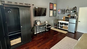 Een woonkamer met een donkere bank, een kamerbreed tapijt, een plafondventilator, een keukenhoek met een magnetron en een aan de muur bevestigde televisie.