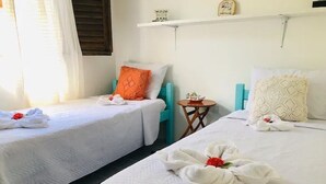 A hotel room with two beds, each with a white bedspread and a towel with a red flower. There is a small round table with a decorative item on it, and a shelf above the beds with a clock and a small plant. - Beach House in Praia dos Carneiros/PE, one of the most beautiful beaches in Brazil (Tamandaré)