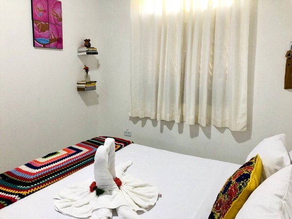 A hotel room with a bed, a colorful blanket, a white towel, a small shelf with books and a vase, and a window with white curtains.