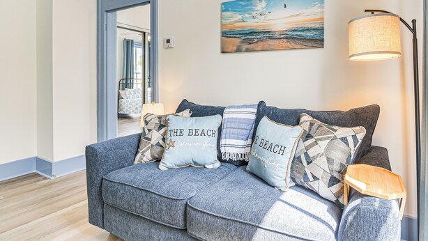 A modern living room with a grey sofa adorned with beach-themed cushions, a painting of a beach scene, and a floor lamp. - Walk to Strand District: Historic Galveston Home (Galveston)