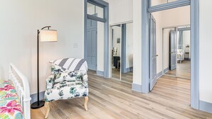 A room with a wooden floor, a white bed with a colorful bedspread, a floral armchair, a floor lamp, and a large mirror reflecting the room. - Walk to Strand District: Historic Galveston Home (Galveston)
