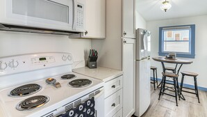 A kitchen with a white stove, microwave, and refrigerator, a dining table with bar stools, and a window with blinds. - 1 Mi to Pleasure Pier: Golden-Age Getaway! (Galveston)