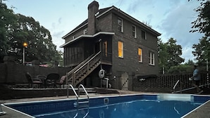 Une piscine rectangulaire dotée d’une terrasse en bois et d’un escalier menant à une maison dont l’extérieur est en briques et qui possède de nombreuses fenêtres.