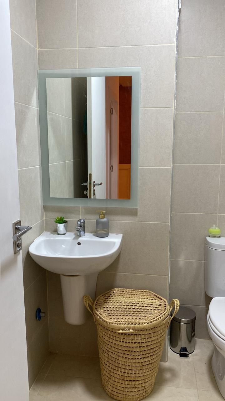Un baño con lavamanos blanco, un espejo, una pequeña planta en maceta, un cubo de basura y un inodoro.