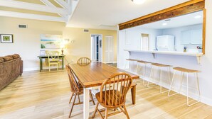 A dining area with a wooden table and chairs, a kitchen with a TV and bar stools, and a living room with a couch and a painting. - Pool Access + Pet Friendly: Condo in Hilton Head (Hilton Head Island)