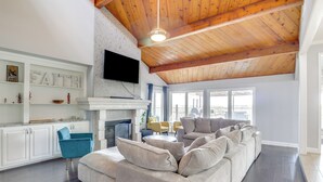 Uma sala de estar ampla com um sofá seccional bege grande, uma poltrona azul e uma lareira. Há uma TV de tela plana montada acima da lareira e um teto de madeira.