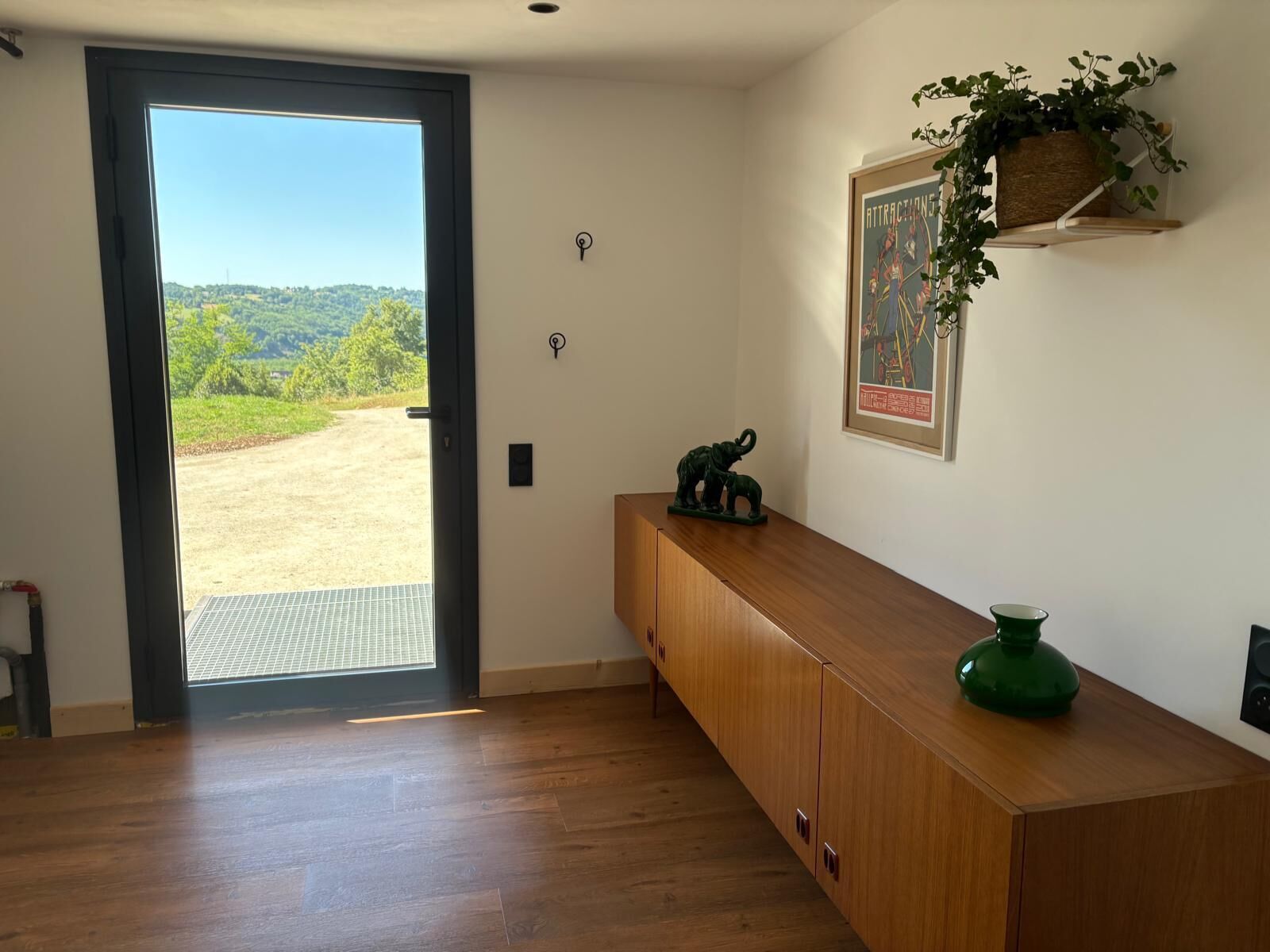 A room with a wooden cabinet, a green vase, a framed picture, and a view of a green landscape through a door.