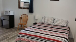 A bedroom with a bed, a chair, a desk with a microwave, and a window with curtains.