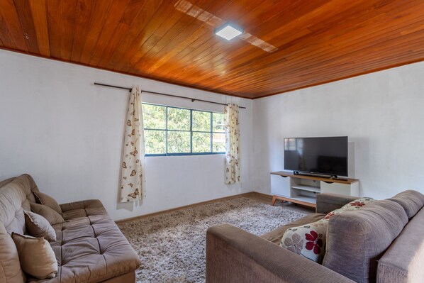 A living room with a wooden ceiling, a large window with curtains, a flat-screen TV, and two sofas.