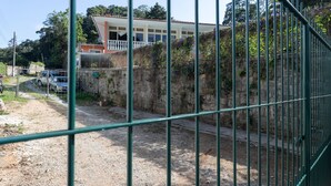 Un área cercada con un edificio al fondo, un camino de grava y coches aparcados.