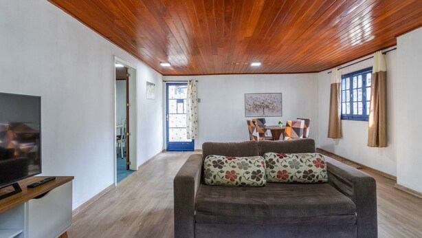 Una sala de estar con techo de madera, un sofá con cojines estampados, un televisor y un comedor al fondo.