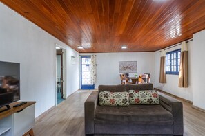 A living room with a wooden ceiling, a sofa with floral cushions, a television, and a dining area in the background.