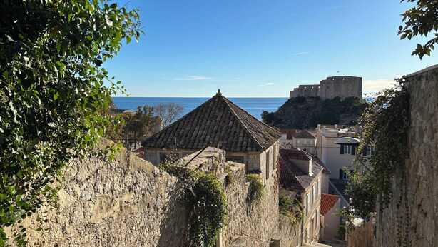 Eine enge Gasse mit Steinbauten, einer steilen Treppe und Blick auf das Meer sowie eine Burg in der Ferne.