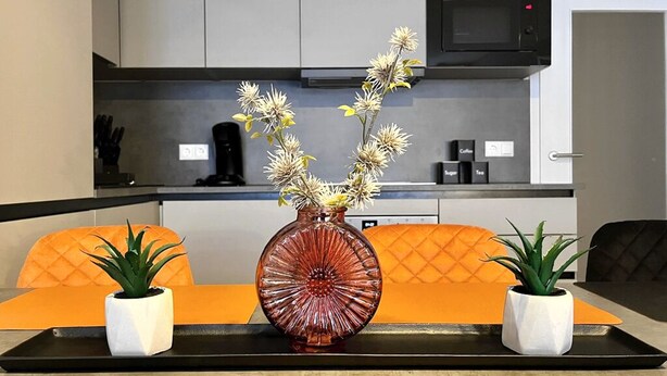 Una cocina moderna con gabinetes grises, microondas y cafetera. Hay un florero con flores sobre una bandeja entre dos sillas naranjas.