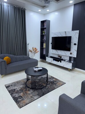 Un salon moderne avec un canapé gris, une table basse noire, un téléviseur fixé sur un mur en marbre et une bibliothèque.