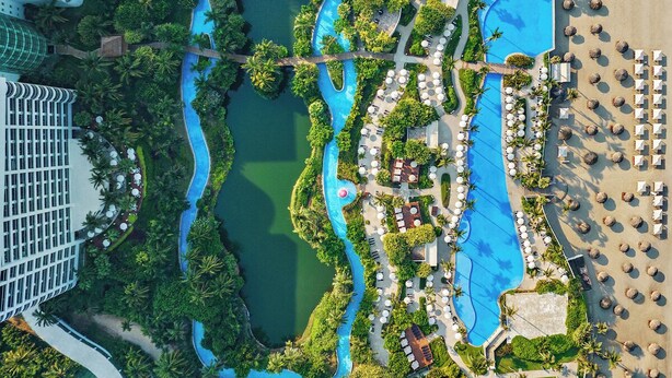 Oversigtsbillede af et resort med flere swimmingpools, frodigt grønt område og en strand med liggestole.